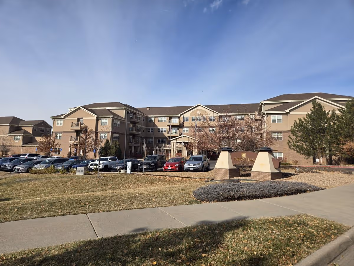 Exterior front view of a multi-story senior living building with a parking lot, landscaping, and a 'Garden Plaza' entrance sign.
