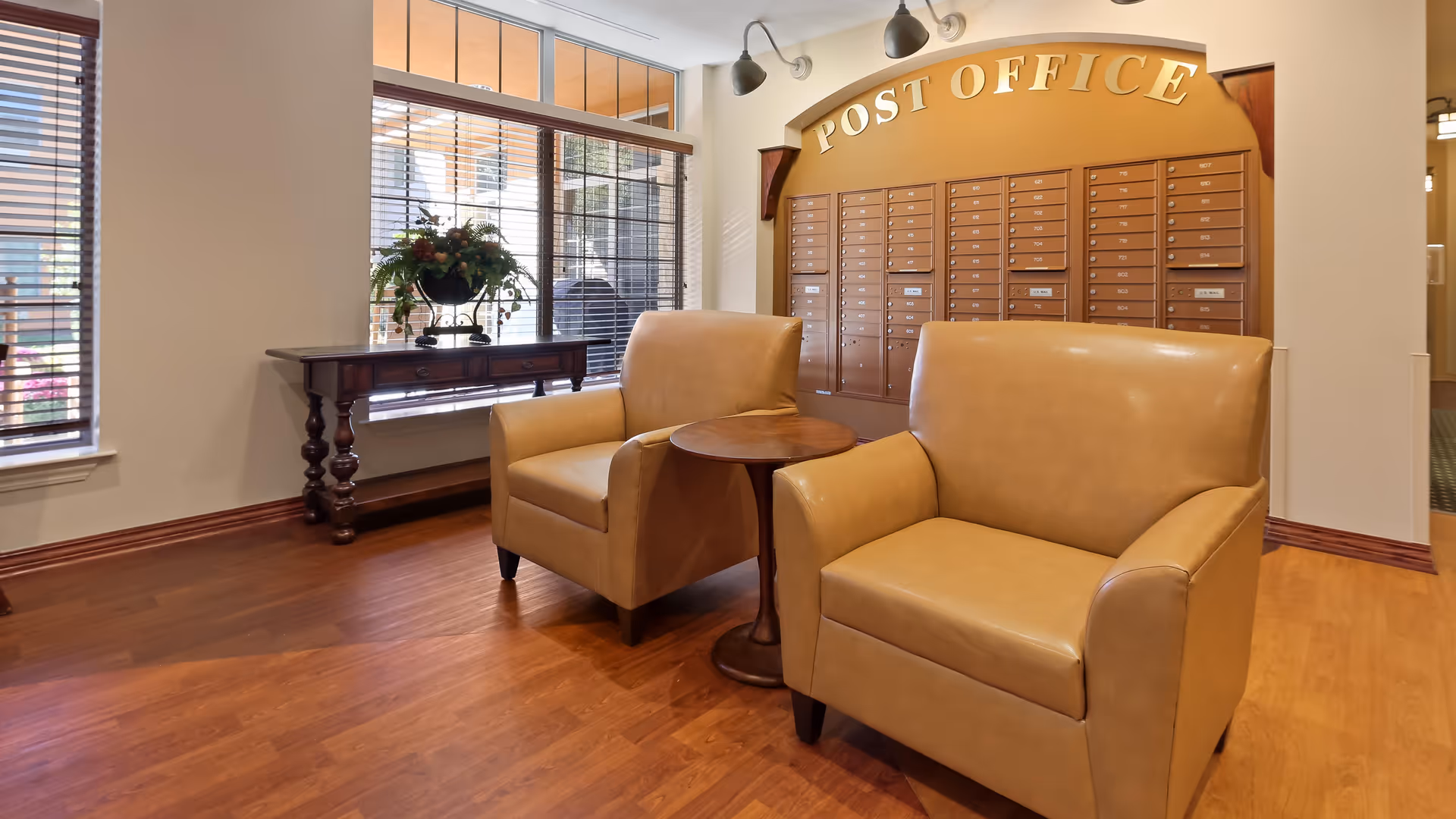 A cozy seating area in a senior living facility with two tan leather armchairs and a small round wooden table between them. Behind the chairs is a wall-mounted post office mailbox unit with multiple mail slots and the words 'POST OFFICE' displayed above. To the left, there is a wooden console table with a decorative flower arrangement in front of large windows with wooden blinds.