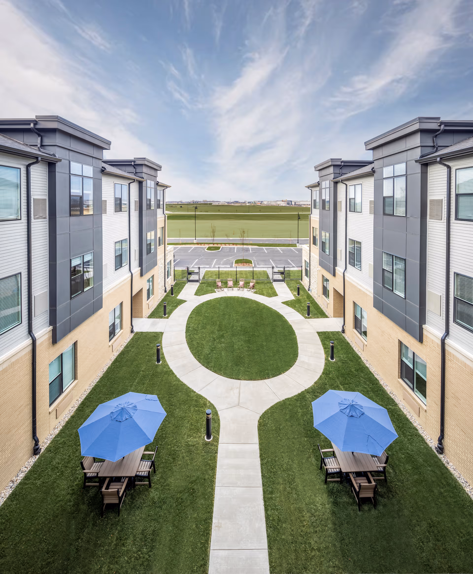 Landscaped central courtyard between two three-story buildings with a circular walkway, green lawn, picnic tables with blue umbrellas, and a view to a parking lot and open field.