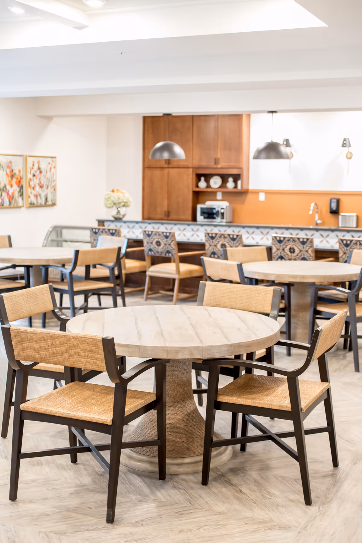 A bright dining area with round wooden tables and chairs featuring woven seats and backs. In the background, there is a kitchen counter with patterned tiles, wooden cabinets, and hanging pendant lights. The walls are decorated with floral paintings and a vase with flowers is placed on a side table.