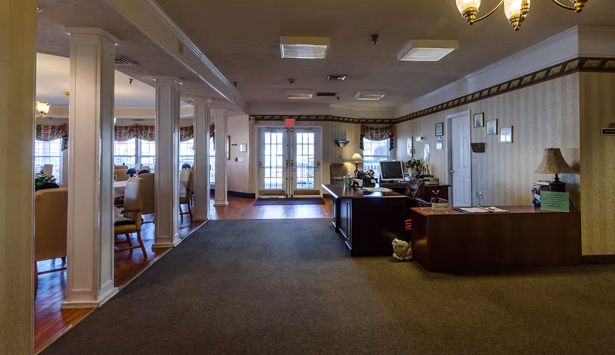 Reception and common area of a senior living facility with a front desk, seating, columns, and dining tables near glass doors.