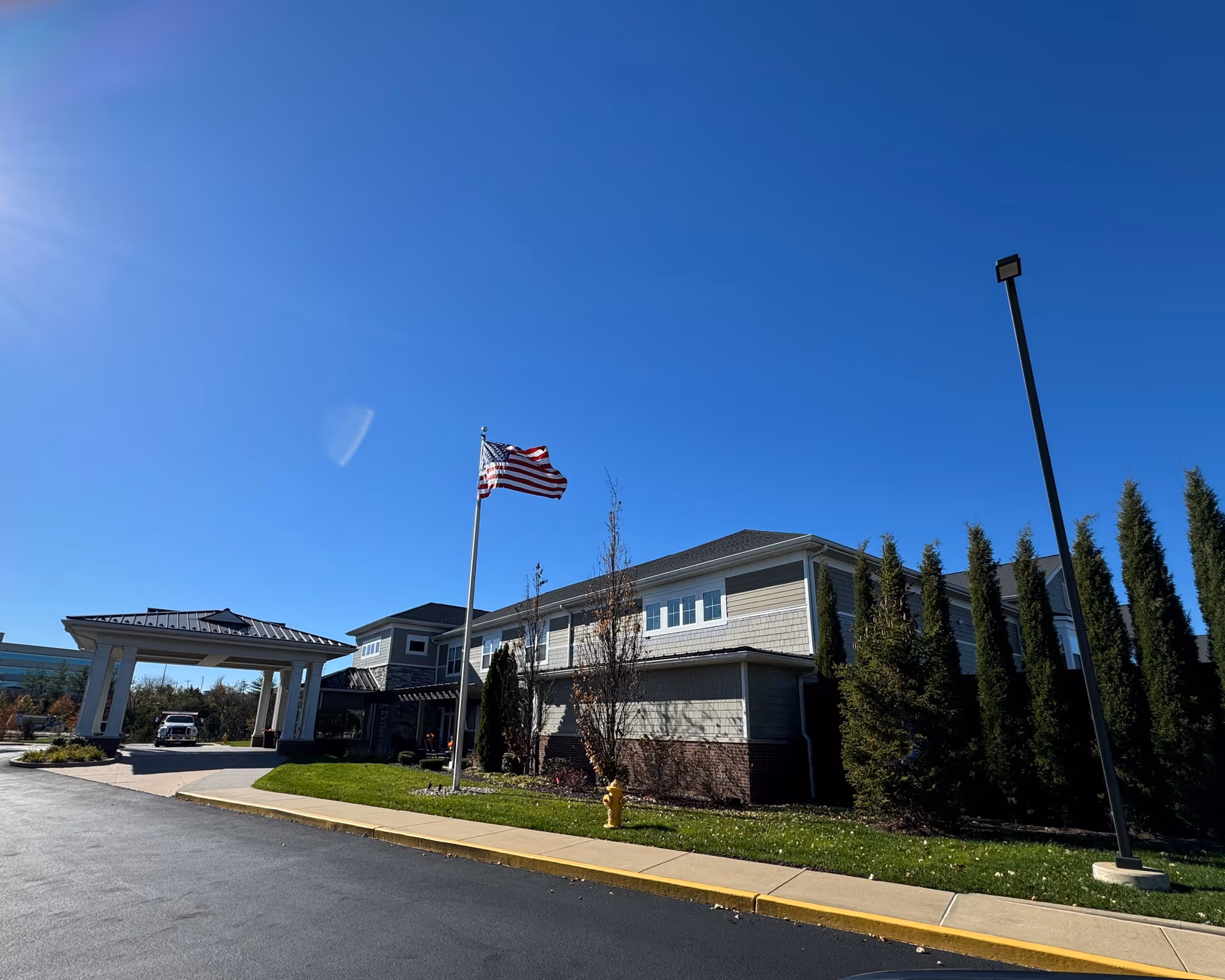 Exterior view of Cedarhurst of Des Peres building under a clear blue sky, featuring an American flag on a flagpole, a covered entrance with a vehicle parked underneath, a row of tall evergreen trees, a street lamp, and a yellow fire hydrant on a grassy area beside the sidewalk.