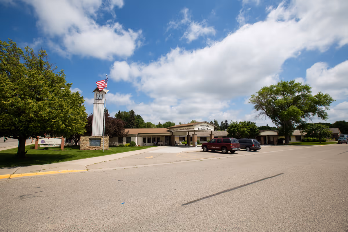 Exterior view of Minnewaska Community Health Services building under a partly cloudy sky. The building is single-story with a covered entrance and a clock tower with an American flag in front. There are several trees and parked cars in the parking lot.