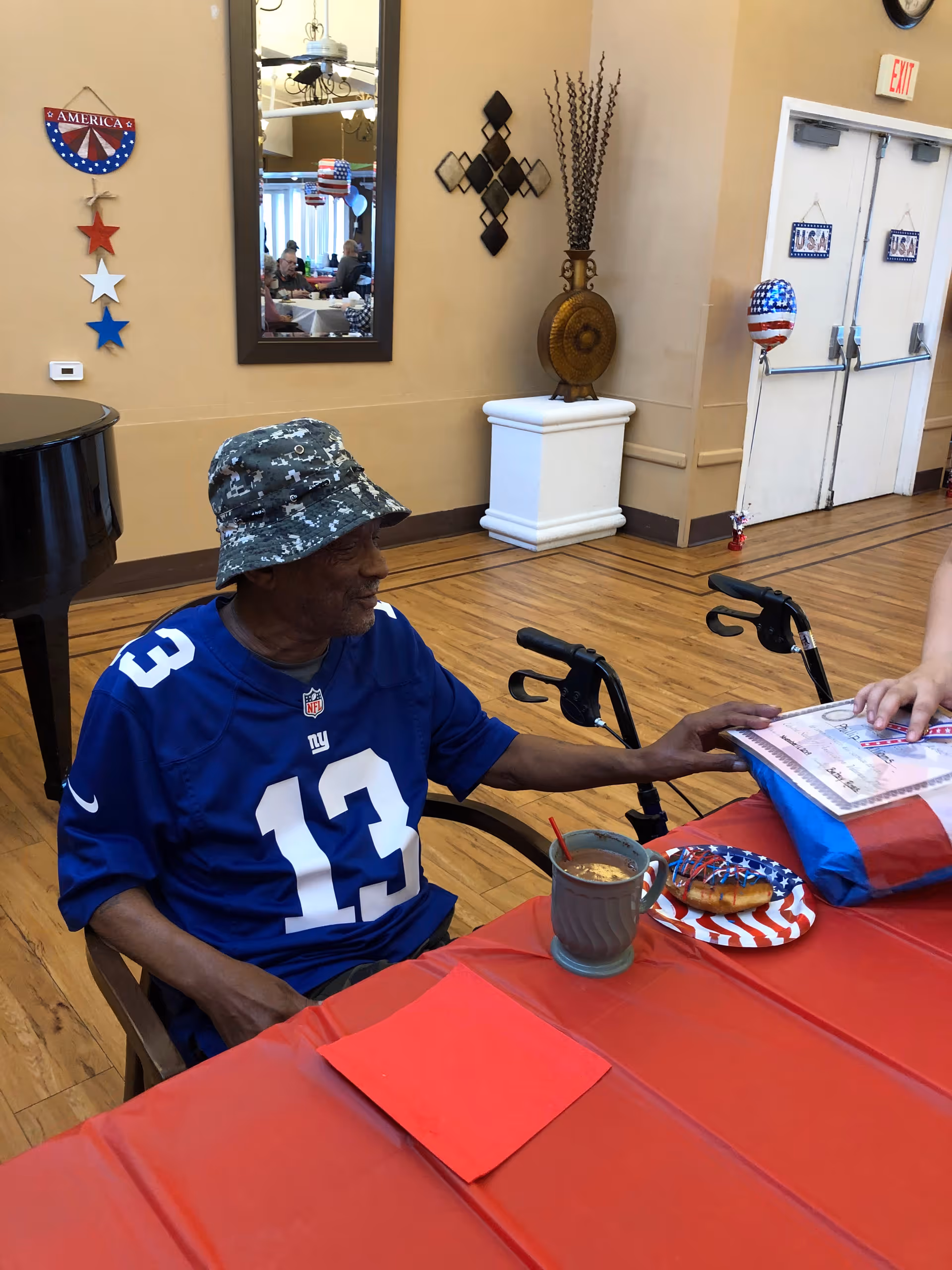 An elderly man wearing a blue New York Giants jersey and a camouflage bucket hat sits at a table covered with a red tablecloth. On the table are a mug with a spoon, a plate with a decorated donut, and a wrapped gift. The room has wooden floors, patriotic decorations on the walls, and a mirror reflecting other people sitting at tables.
