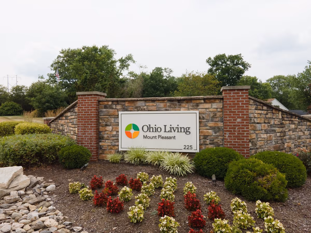 Stone and brick entrance sign for Ohio Living Mount Pleasant with landscaping including bushes and red and white flowers in front, and trees in the background under a cloudy sky.