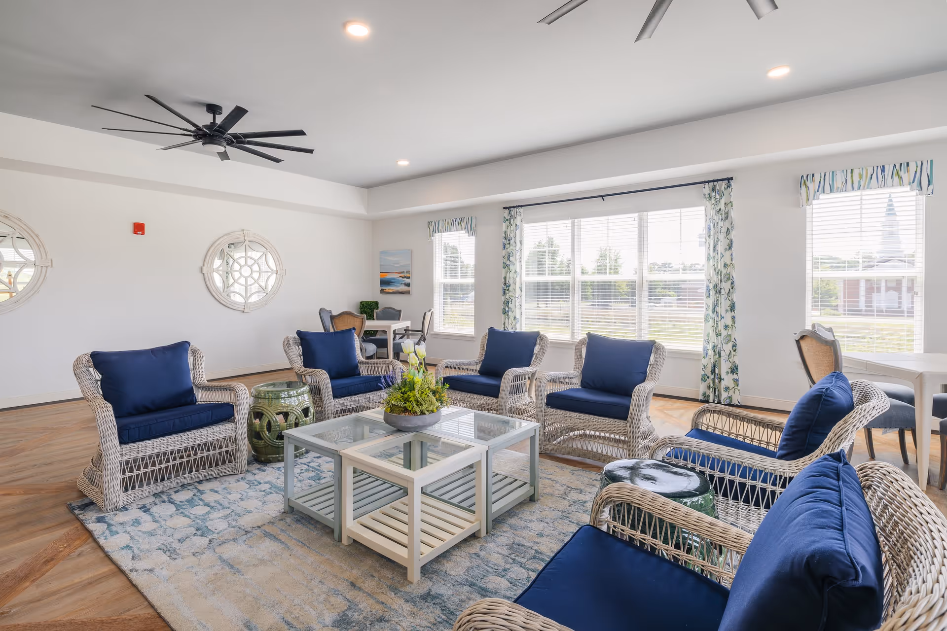 Bright and spacious living room with wicker armchairs featuring navy blue cushions arranged around a glass coffee table on a patterned rug. Large windows with floral curtains allow natural light to fill the room. The walls are white with two decorative round mirrors, and a large black ceiling fan is mounted on the ceiling. Additional seating and a small table are visible in the background.