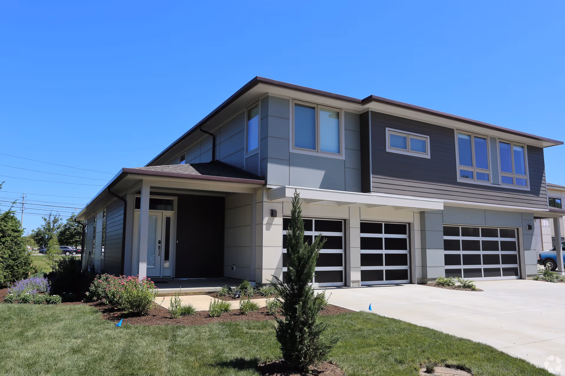 Modern two-story residential building with a well-maintained lawn and small shrubs in front, featuring large windows and three garage doors under a clear blue sky.