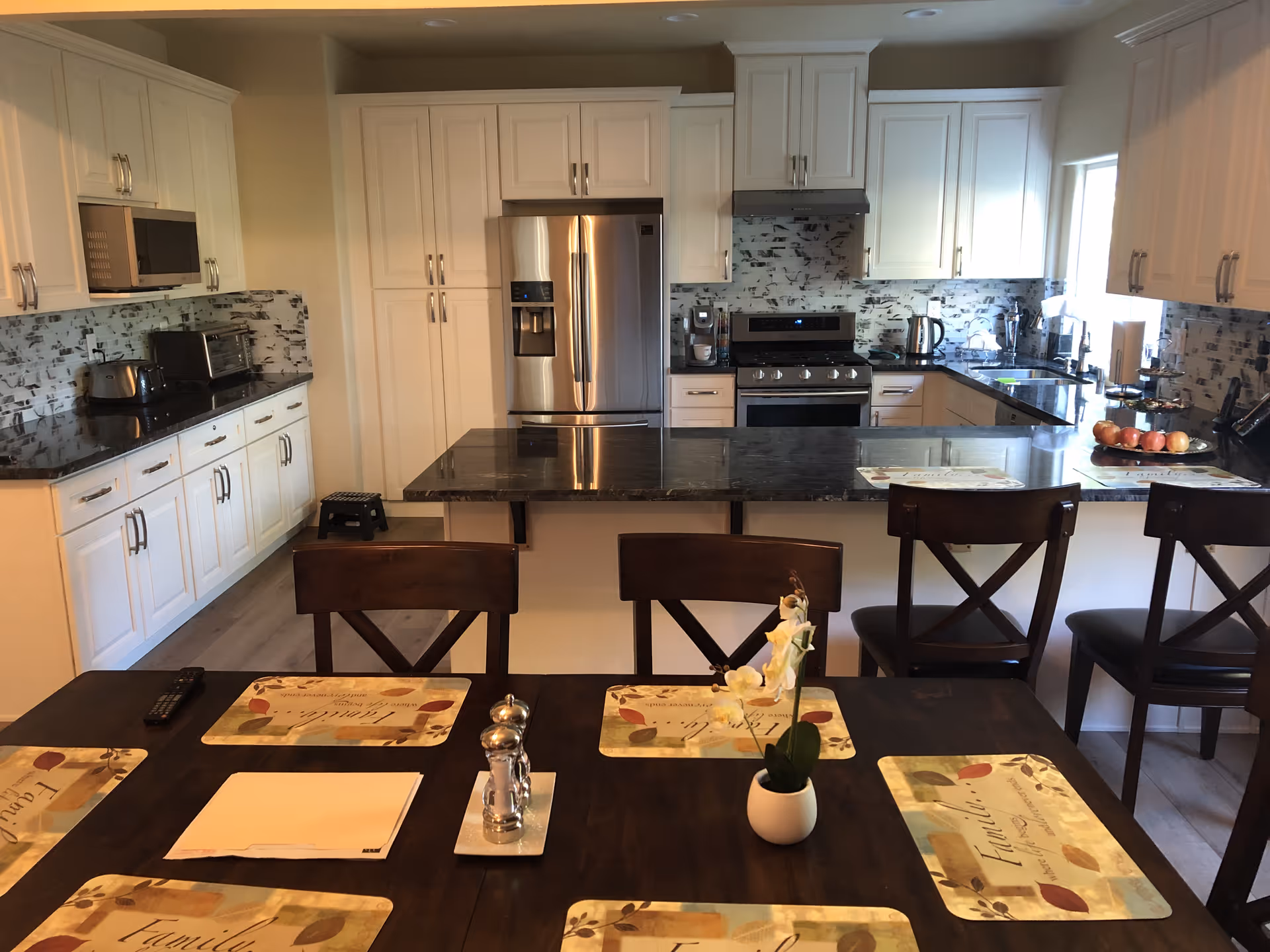 A modern kitchen with white cabinets, a stainless steel refrigerator, stove, microwave, and black granite countertops. There is a kitchen island with three wooden chairs and a dining table in the foreground set with placemats, a small potted plant, salt and pepper shakers, and a remote control.