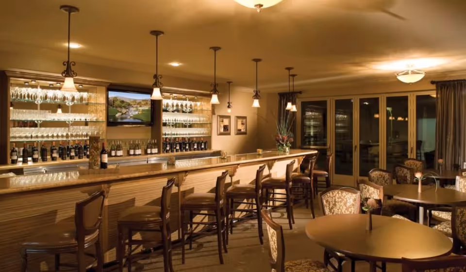 Interior view of a warmly lit bar area with a long counter, several high wooden chairs, shelves stocked with wine bottles and glasses, a mounted TV screen, and round tables with upholstered chairs arranged nearby.