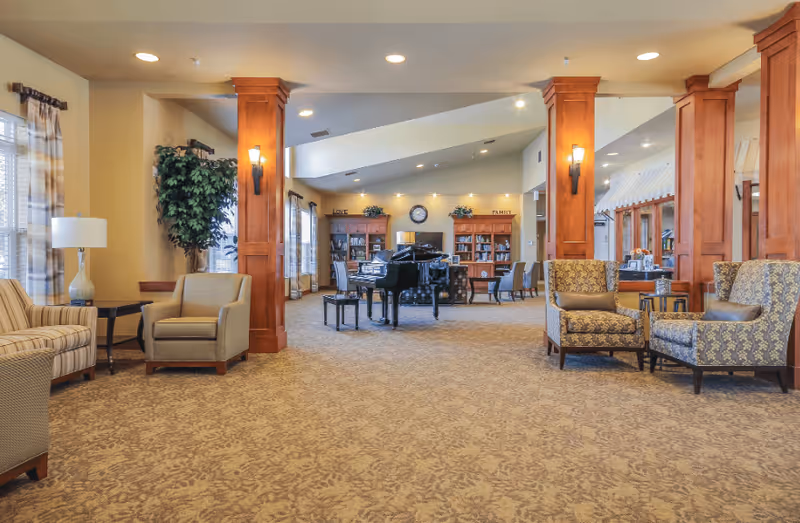 A spacious and well-lit living room area in an assisted living facility featuring comfortable armchairs and sofas arranged around wooden columns. In the background, there is a black grand piano with a bench, bookshelves filled with books, and a clock on the wall. The room has large windows with curtains, carpeted floors, and warm lighting from wall sconces and ceiling lights.