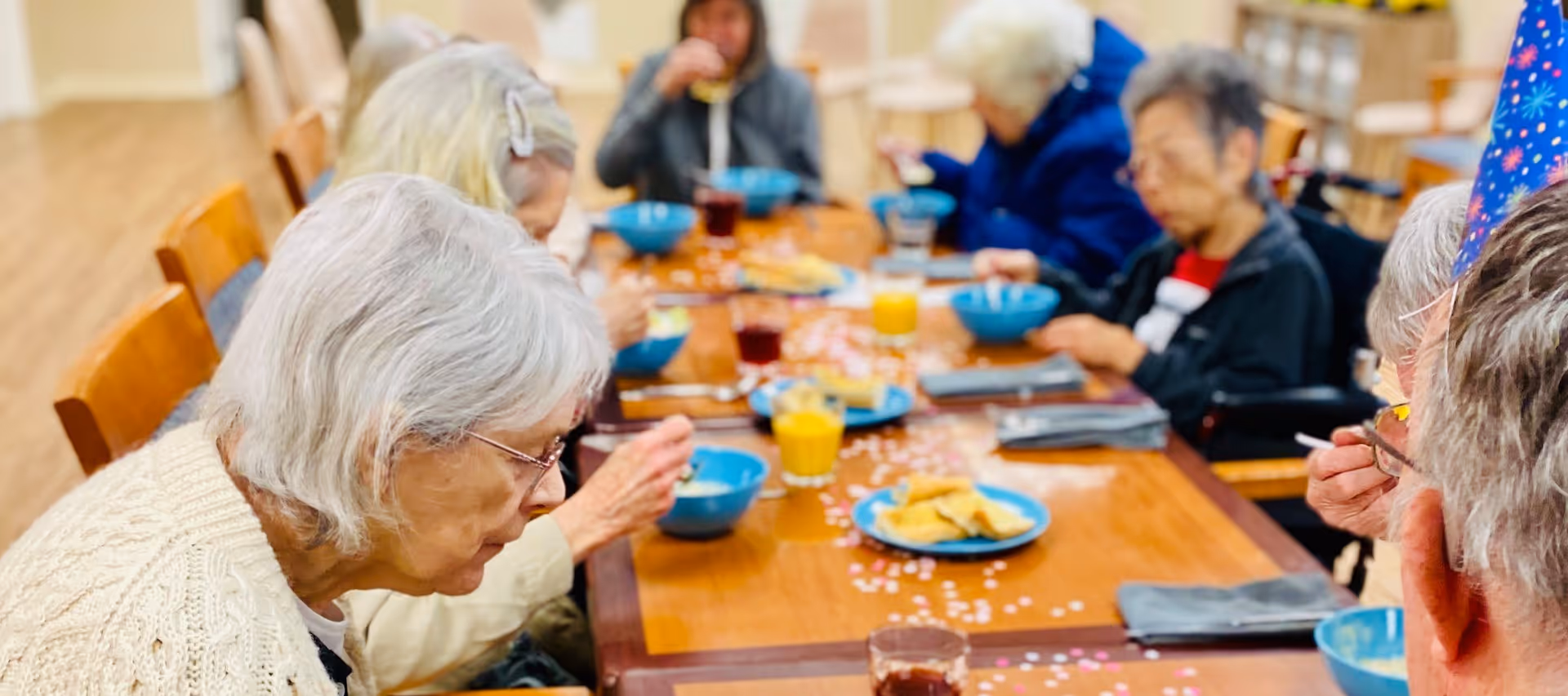 A group of elderly people sitting around a wooden dining table eating from blue bowls. The table is decorated with pink and white confetti and has plates of food and glasses of juice and other beverages. The setting appears to be a communal dining area in a senior living facility.
