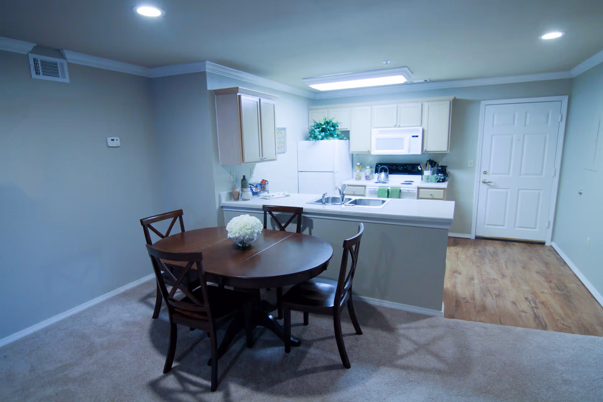 Open-plan kitchen and dining area with a round wooden table and four chairs in front of a white kitchenette and island sink.