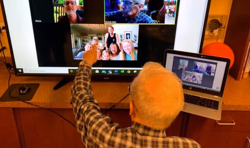 An elderly man with white hair and glasses is sitting in front of a large screen and a laptop, participating in a video call with multiple people visible on the screens. He is pointing at the large screen showing several video call participants.