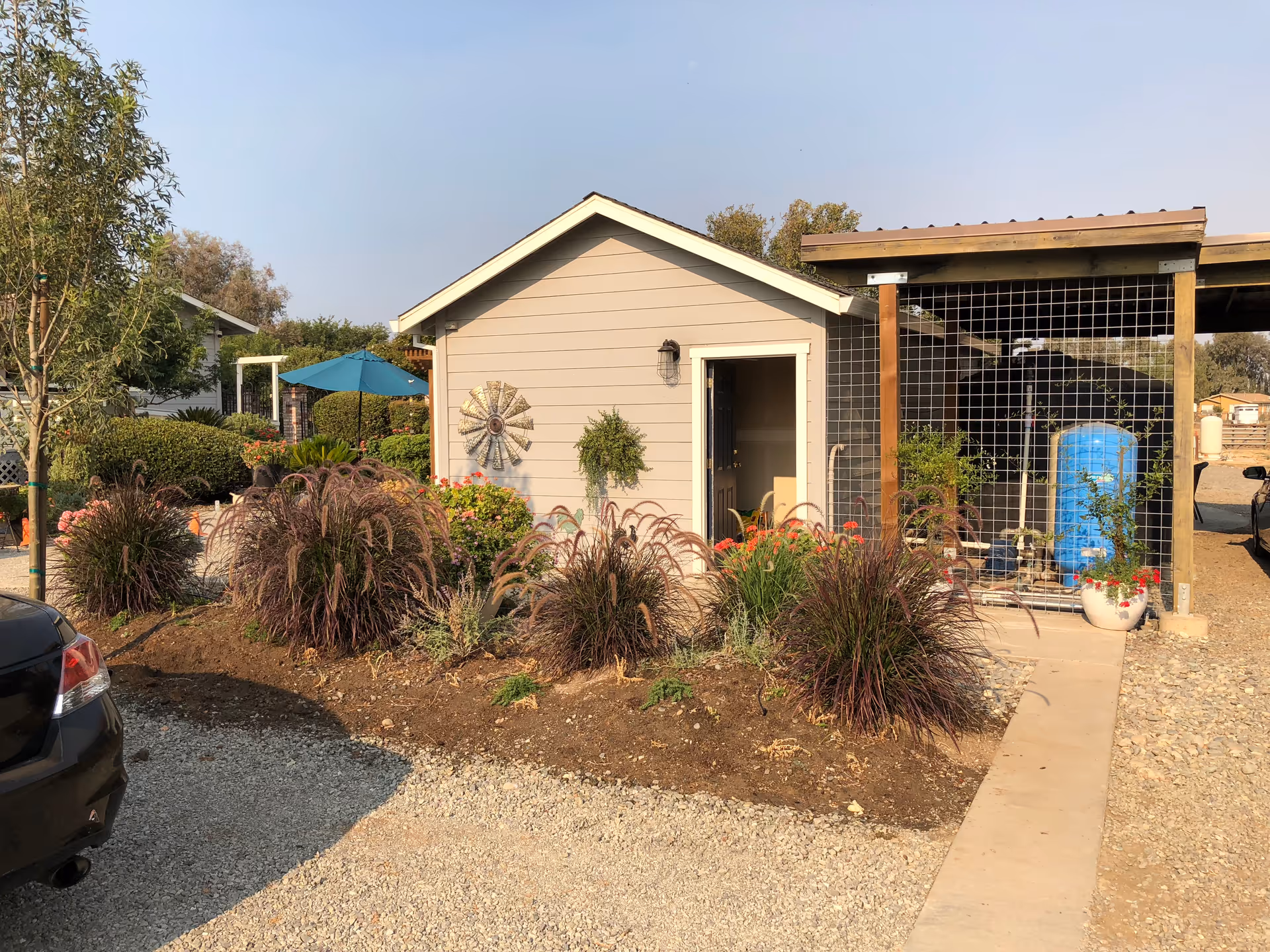 Exterior view of a small beige building with a gable roof, surrounded by landscaped plants and bushes. There is a decorative metal windmill on the building's wall and a hanging plant near the door. To the right, there is a covered area with a wire mesh enclosure containing a blue water tank and some potted plants. A black car is partially visible on the left side of the image, and a gravel driveway leads up to the building. The sky is clear and blue.