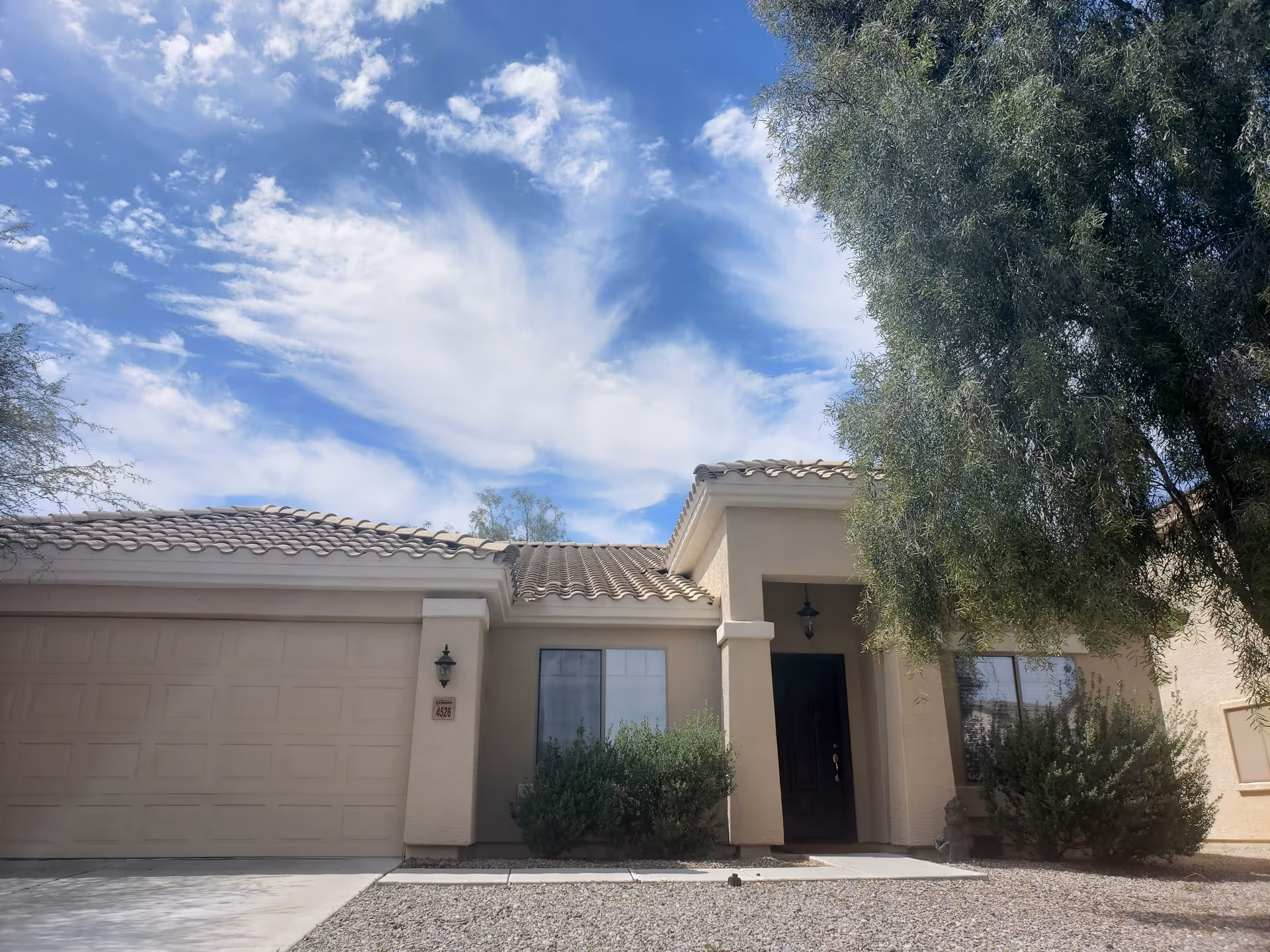Front exterior view of a single-story beige stucco building with a tiled roof, a garage door on the left, a black front door under a small covered porch, and bushes in front of the windows. A large tree is on the right side, and the sky is partly cloudy with blue patches.