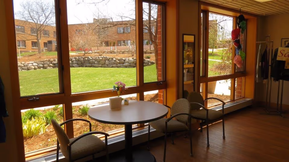 A small seating area inside a building with a round table and three chairs near large windows. Outside the windows, there is a green lawn, a stone retaining wall, and a brick building in the background. Inside, there is a rack with hanging items and a sign partially visible.