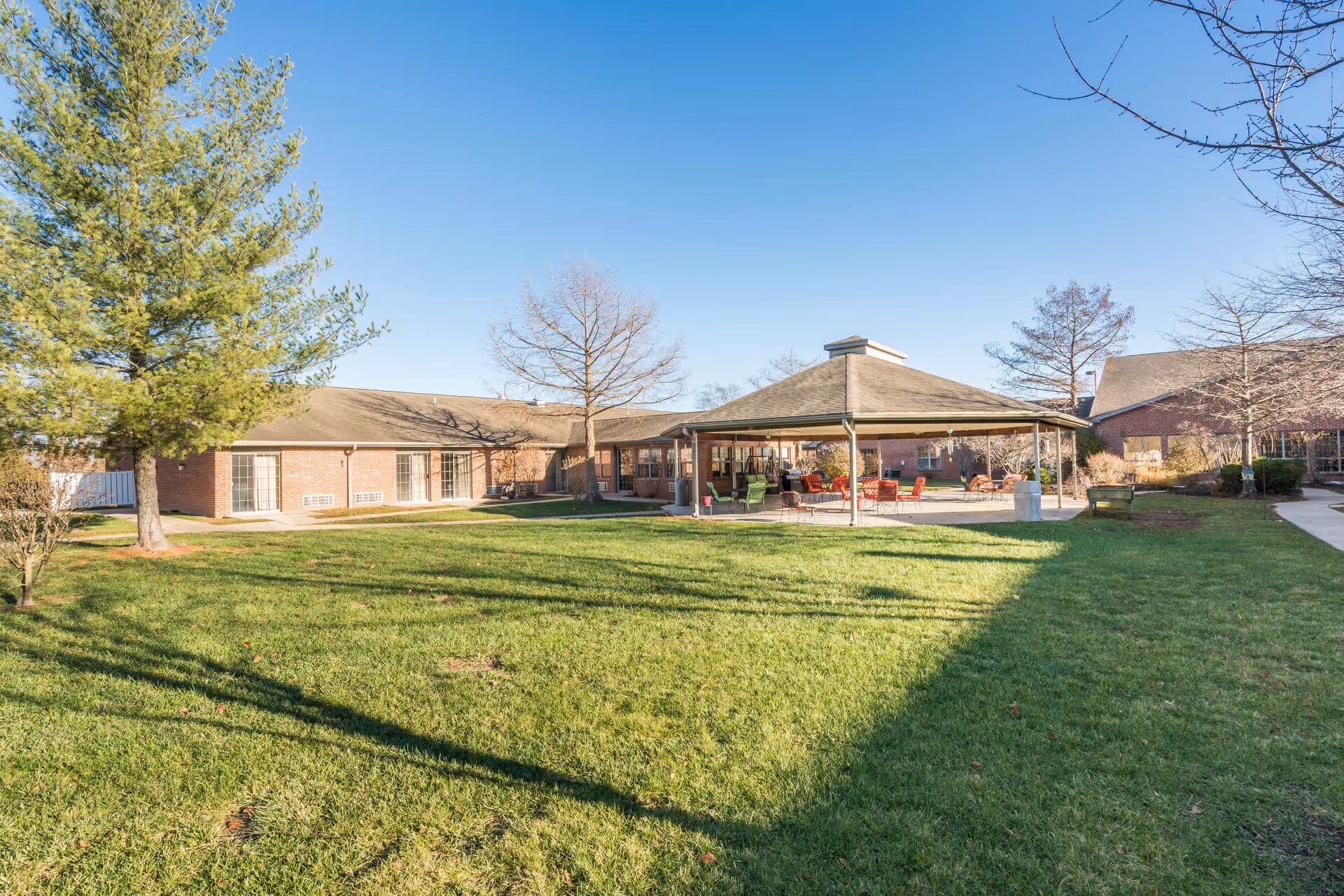 Outdoor view of a senior living facility with a large grassy lawn, several trees, and a covered patio area with chairs and tables. The building is made of brick and has multiple windows. The sky is clear and blue.