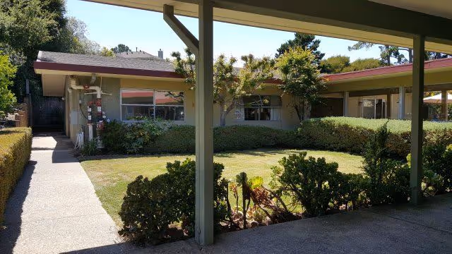 View of a single-story building with a covered walkway in the foreground, surrounded by a well-maintained garden with bushes and small trees under a clear sky.