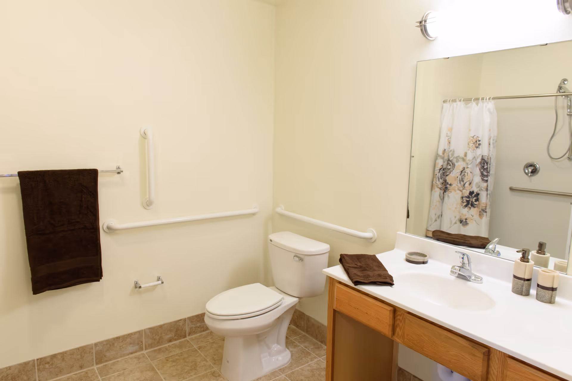 A clean and accessible bathroom with beige tiled floor and cream-colored walls. The bathroom features a white toilet with grab bars on the walls around it, a wooden vanity with a white countertop and sink, a large mirror above the sink, a brown towel on the countertop, and a matching brown towel hanging on a towel rack. A shower with a floral-patterned curtain is visible in the mirror reflection.