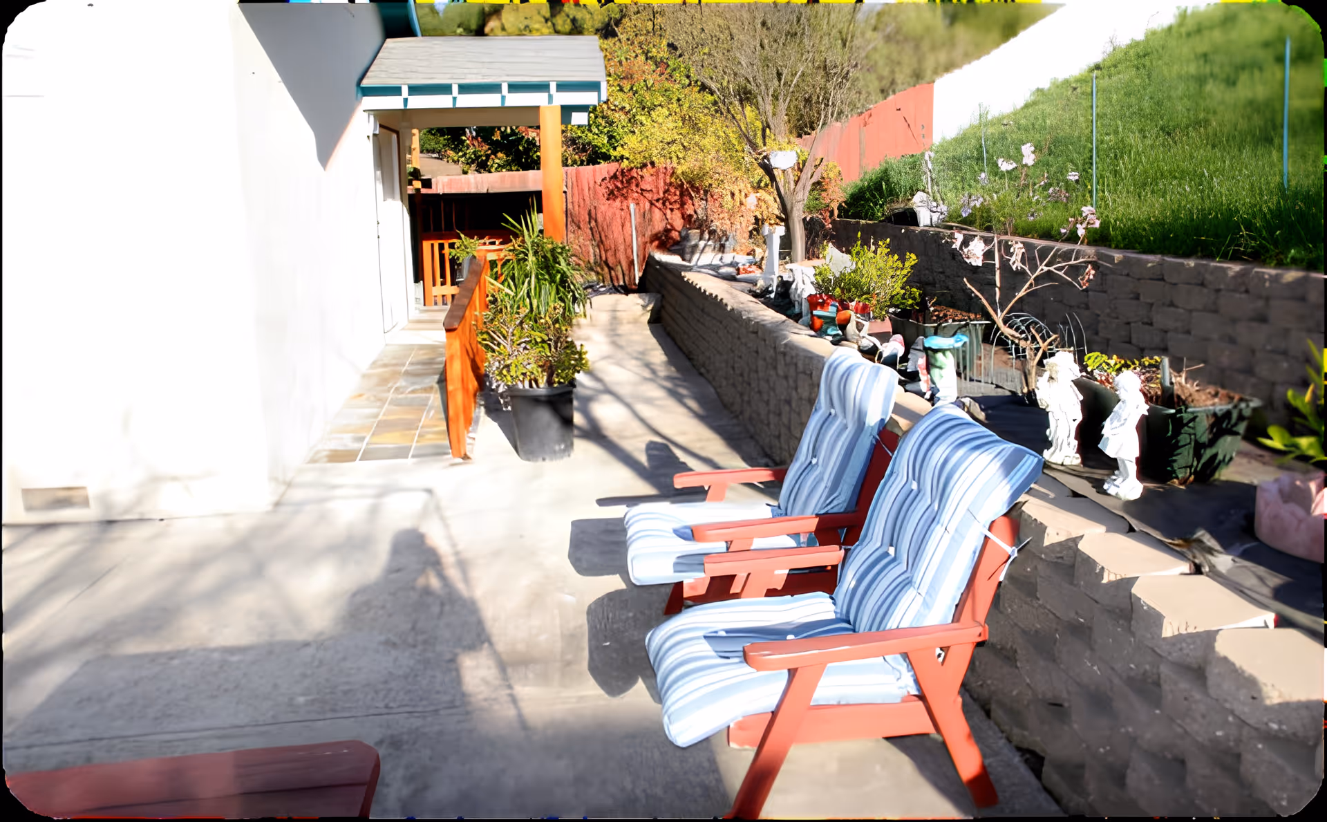 Outdoor patio area with two wooden chairs featuring blue and white striped cushions. The patio is adjacent to a white building with a tiled walkway and a small covered porch. There are various potted plants and garden decorations along a stone retaining wall, with greenery and trees in the background.