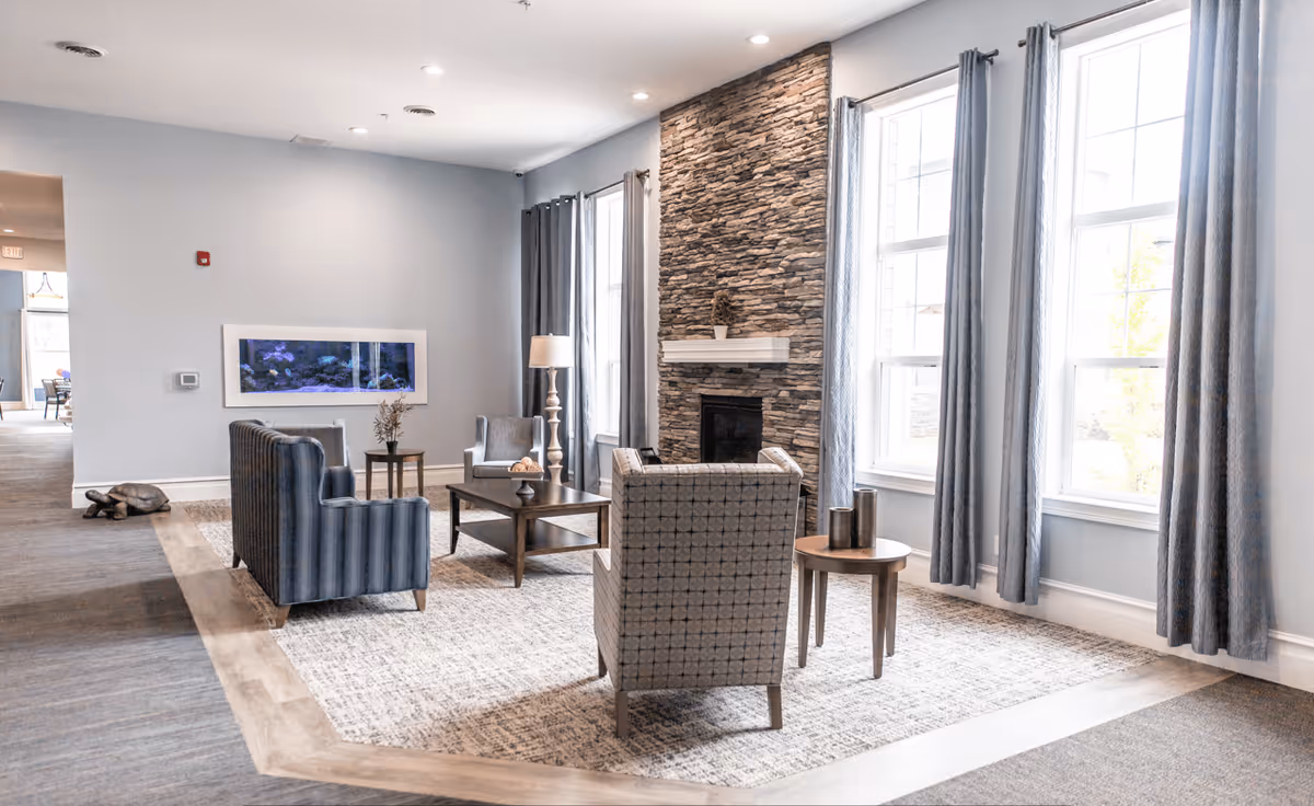 A bright and spacious living room area with large windows covered by blue curtains, a stone fireplace, a blue striped sofa, two patterned armchairs, a wooden coffee table, and side tables with decorative items. The room has light gray walls and a mix of carpet and wood flooring.
