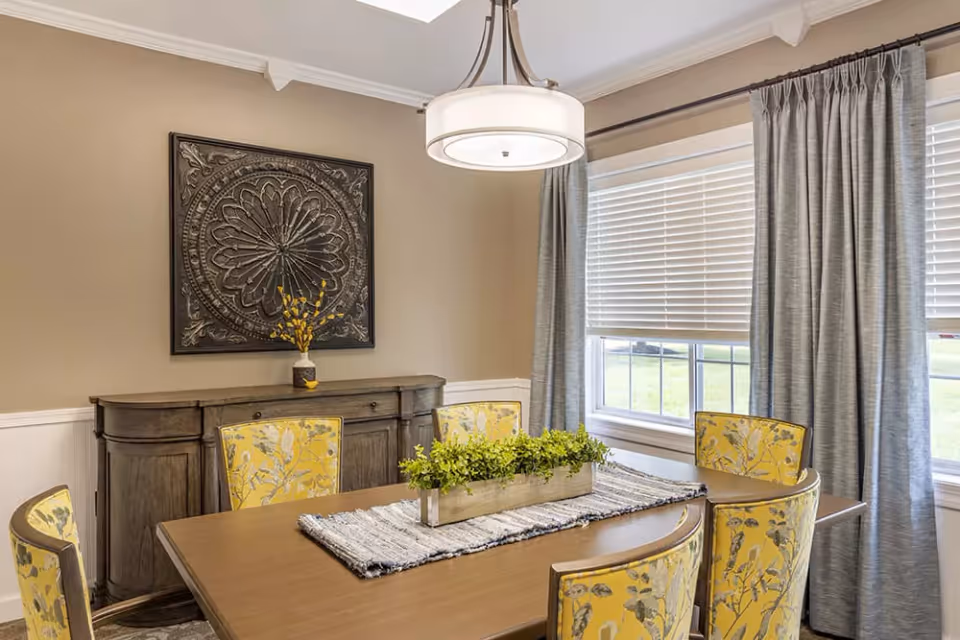 A dining room with a wooden table surrounded by six yellow floral-patterned chairs. On the table is a rectangular planter with green plants and a woven table runner. Behind the table is a wooden sideboard with a decorative vase holding yellow flowers and a large ornate wall art piece above it. The room has beige walls, white trim, a window with white blinds and gray curtains, and a modern circular ceiling light fixture.