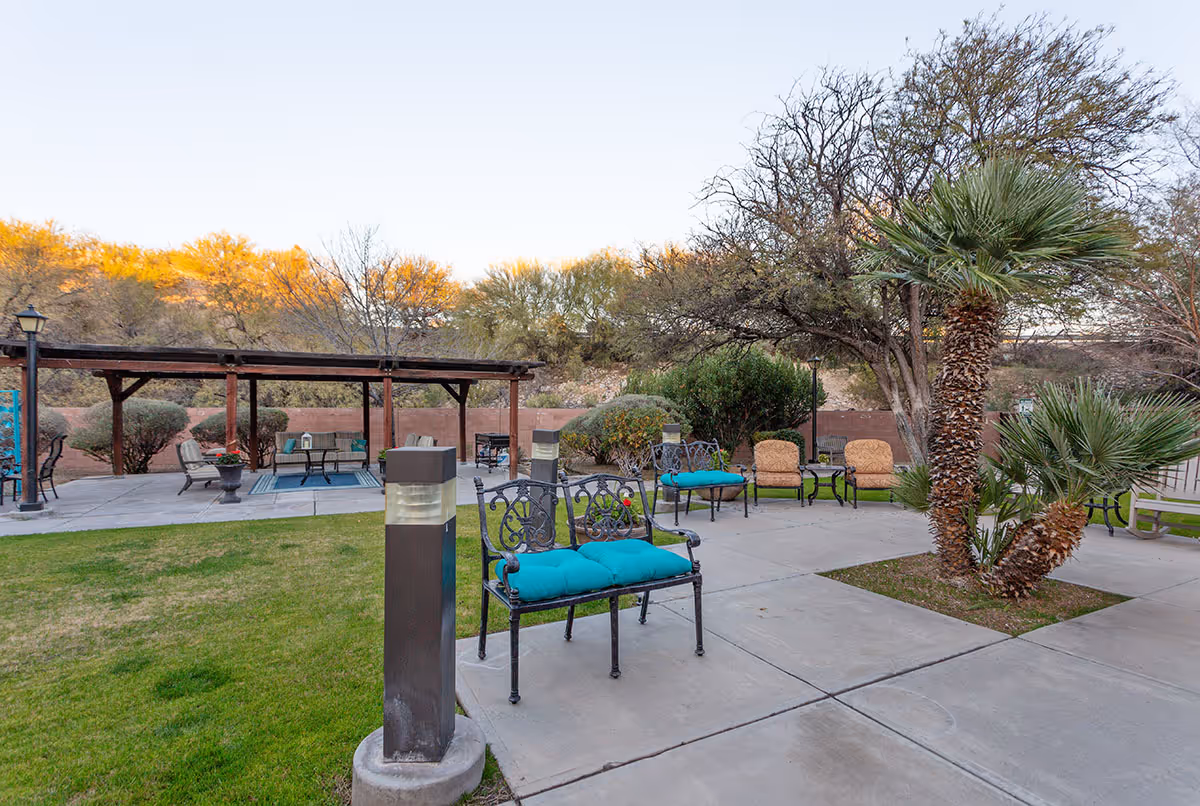 Outdoor courtyard with metal benches and turquoise cushions, chairs under a pergola, palm trees, and paved walkways.