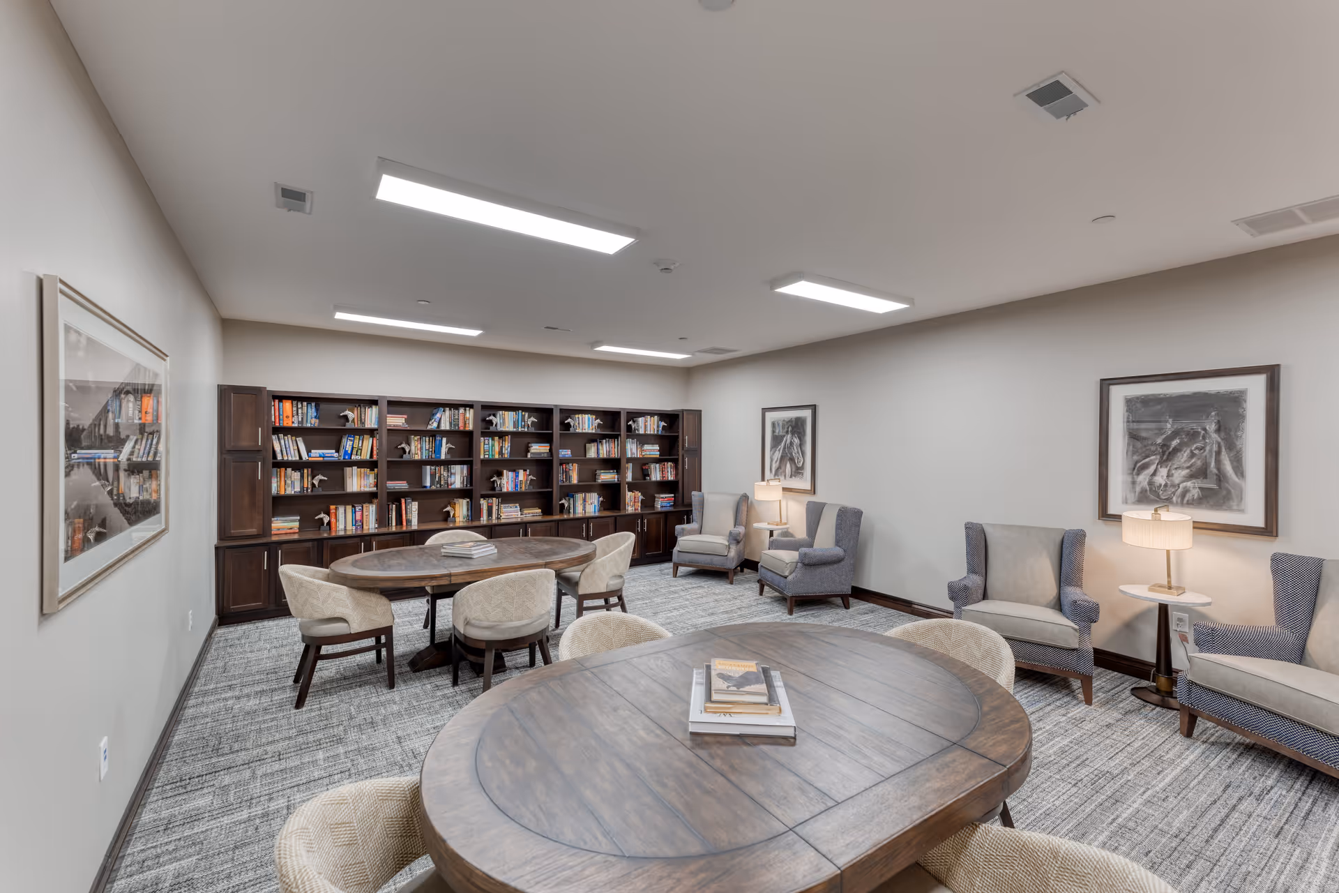 A bright communal reading room with round wooden tables, upholstered chairs, bookshelves and armchairs.
