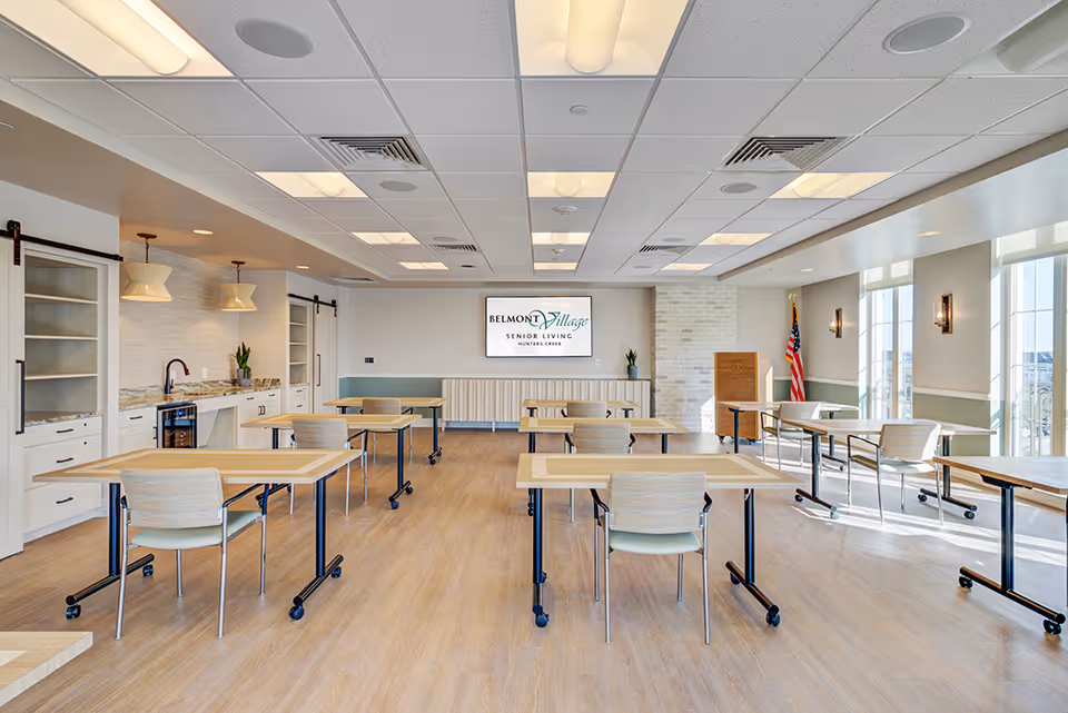 Spacious, well-lit communal dining/activity room with several tables and chairs, a kitchenette on the left, and a sign that reads Belmont Village Senior Living on the back wall.