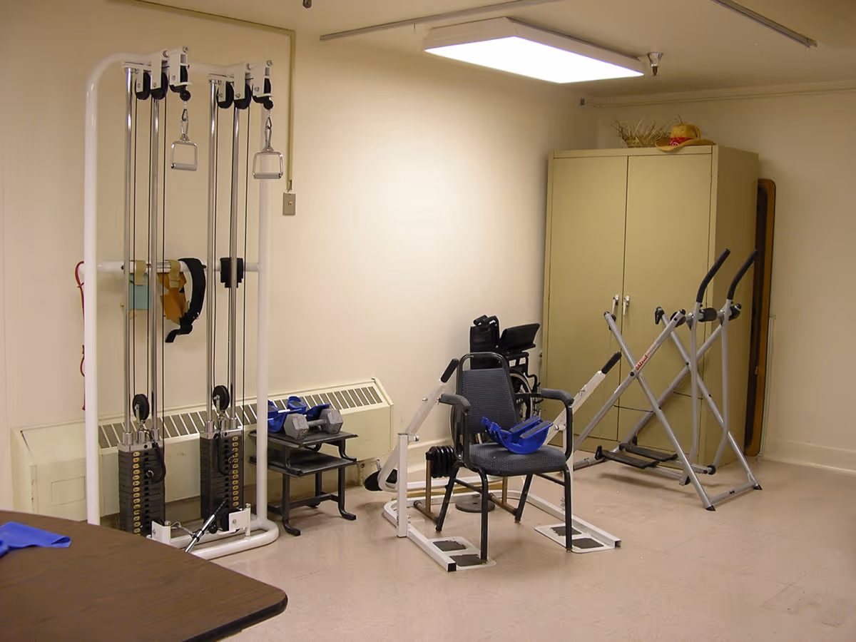 A small exercise room with various fitness equipment including a weight machine, a chair with attached leg exercise apparatus, a wheelchair, dumbbells on a small table, and two elliptical machines. There is a beige cabinet in the corner and a fluorescent light on the ceiling.