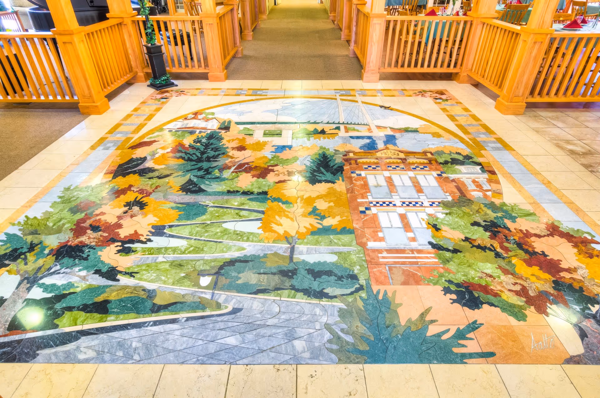 A colorful tiled floor mural depicting a scenic landscape with trees, a winding path, a bridge, and a building, located inside a facility with wooden railings and carpeted flooring.
