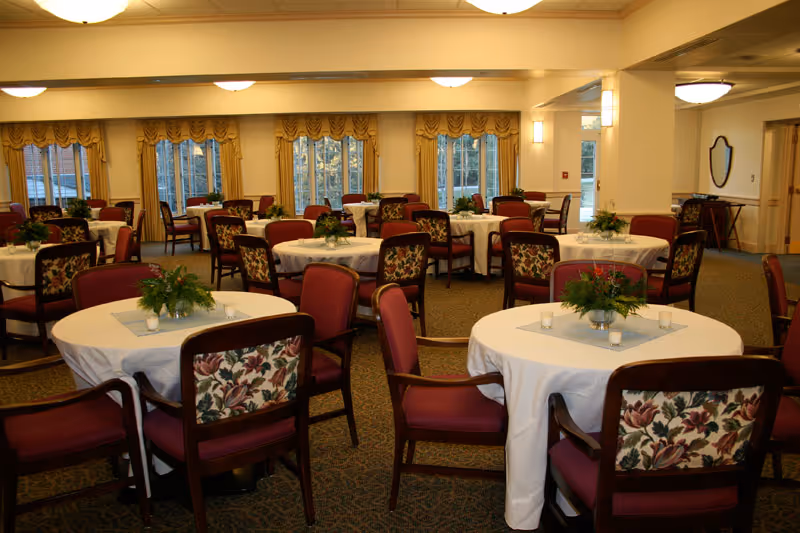 A spacious dining room with multiple round tables covered with white tablecloths. Each table is surrounded by wooden chairs with floral-patterned backs and red cushions. Small green plants and candles are placed as centerpieces on the tables. Large windows with yellow curtains allow natural light to fill the room.