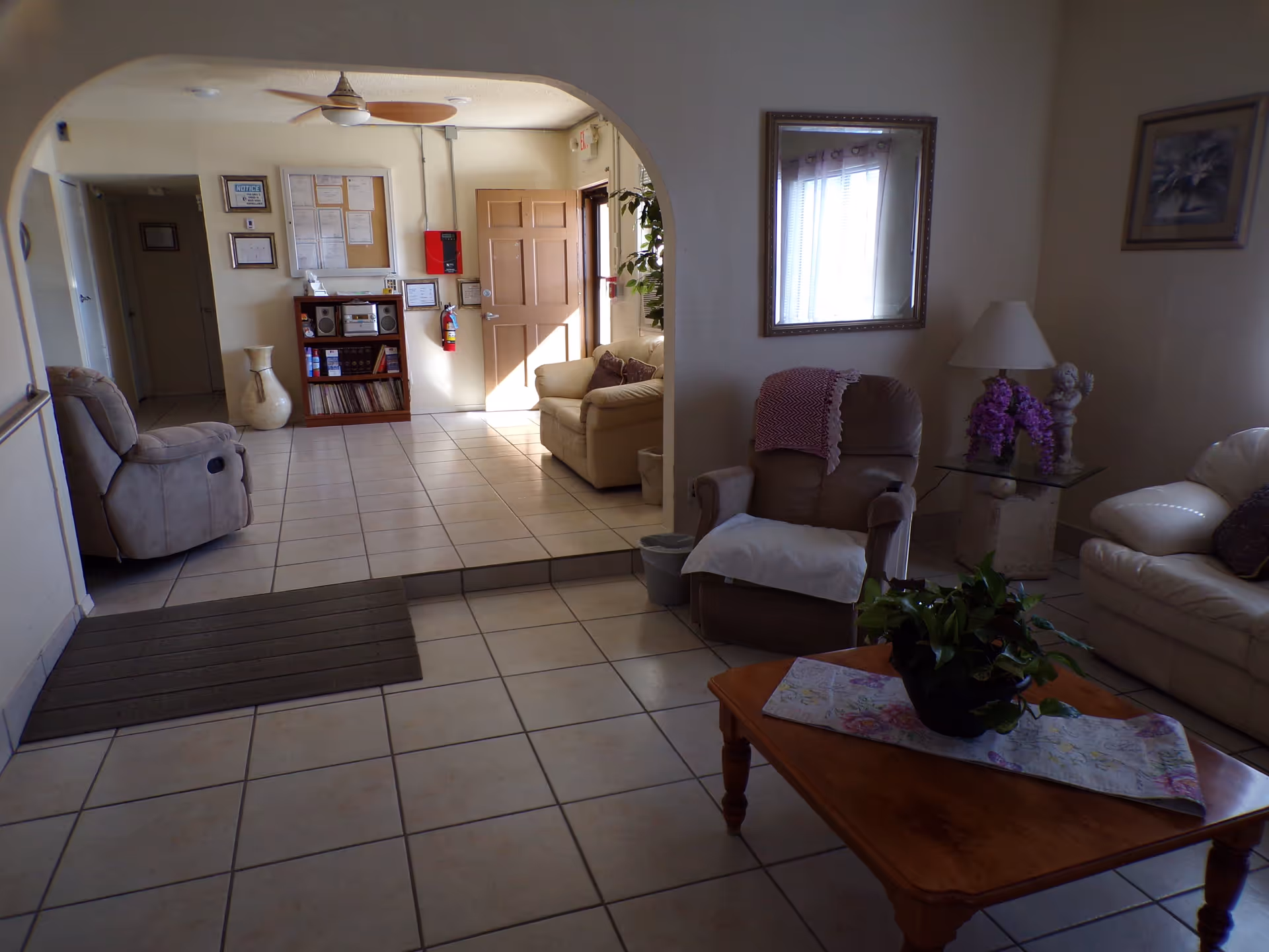 A cozy living room area with beige tiled floors, a brown recliner chair with a white cloth draped over it, a beige sofa, a wooden coffee table with a plant and a floral table runner, and a glass side table with a lamp and purple flowers. The room has a large mirror on the wall and a framed picture. In the background, there is an open doorway leading to a small lobby area with another beige sofa, a wooden bookshelf with books and a stereo, a fire extinguisher, and various notices on the wall.