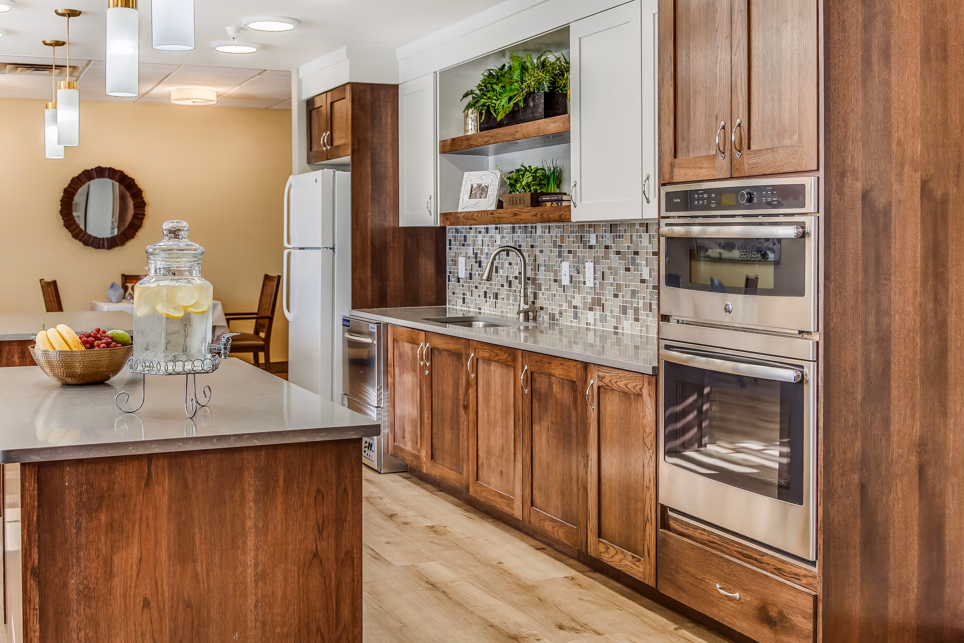 Modern kitchen with wooden cabinets, a double oven, a refrigerator, and a countertop island with a glass container of lemon water and a bowl of fruit. The backsplash features mosaic tiles, and there are plants and decorative items on open shelves above the sink. In the background, a dining area with chairs and a round mirror on the wall is visible.