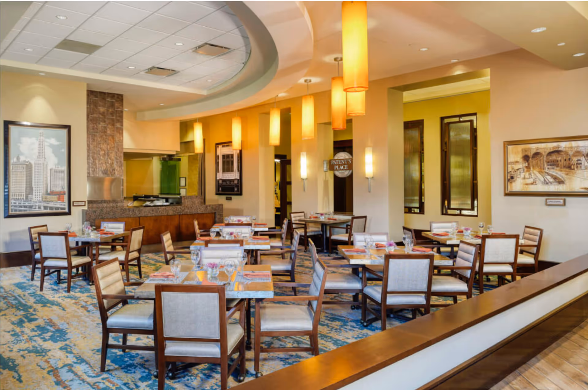A well-lit dining room with multiple tables and chairs arranged neatly. The tables are set with glasses, napkins, and small flower vases. The room features a patterned carpet, warm yellow walls, hanging cylindrical pendant lights, and framed artwork on the walls. There is a counter area in the background and a sign that reads 'PATIENT'S PLACE'.