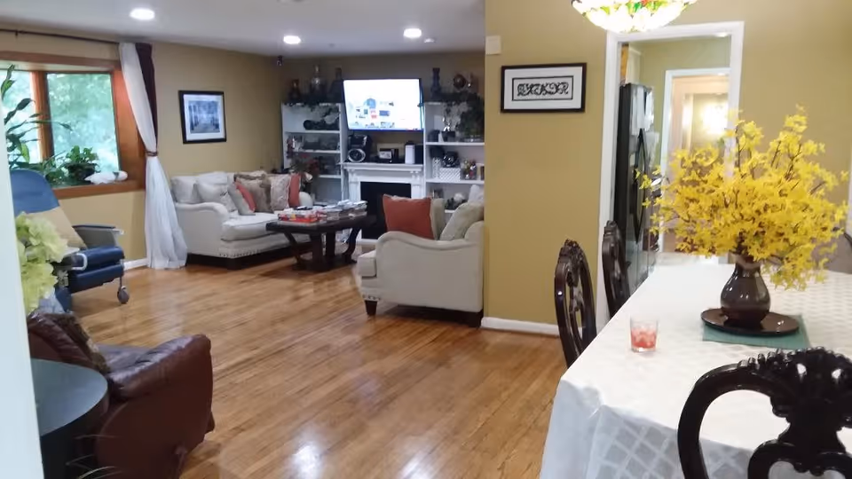 A cozy living room and dining area in a senior living facility with wooden floors, beige walls, and comfortable seating including sofas and chairs. A TV is mounted above a white fireplace with shelves on either side. The dining table is covered with a white tablecloth and decorated with a vase of yellow flowers. Large windows with curtains allow natural light into the space.