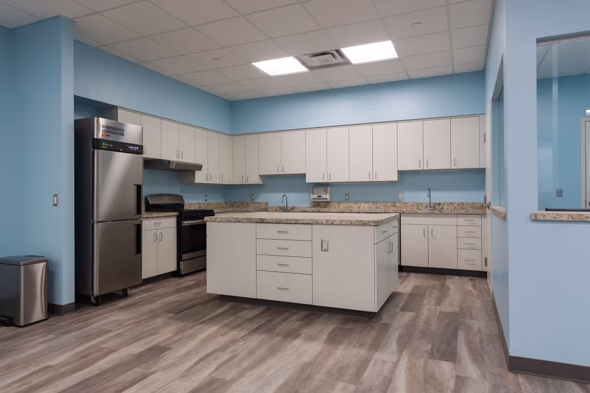 A modern kitchen with light blue walls, light wood cabinets, and granite countertops. The kitchen features a large central island with drawers, a stainless steel refrigerator, a stove with a hood, and two sinks. The floor is covered with wood-patterned vinyl planks, and there is a trash can in the corner.