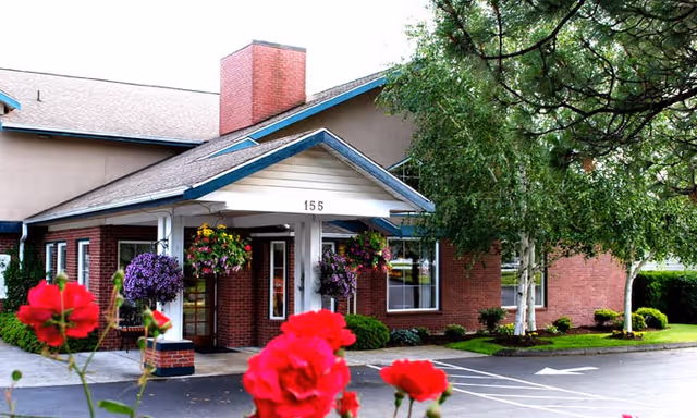 Exterior view of a senior living facility building with a covered entrance marked 155, surrounded by hanging flower baskets, red flowers in the foreground, and trees on the side.