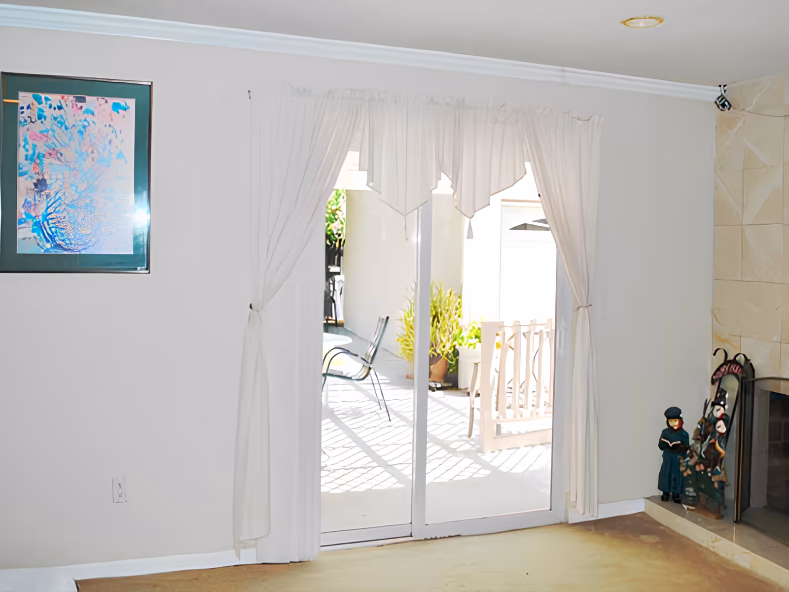 Interior view of a room with a sliding glass door covered by white curtains leading to an outdoor patio area with chairs and plants. On the right side, there is a fireplace with decorative figurines on the hearth. A framed abstract artwork is hanging on the left wall.