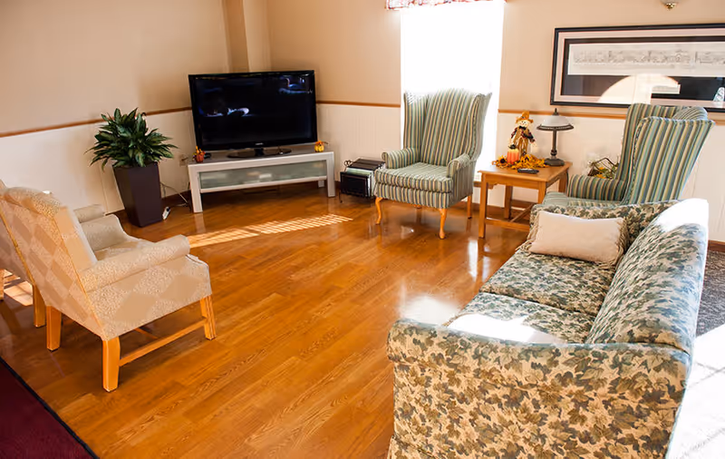 A cozy living room with wooden flooring, featuring a floral patterned sofa with a beige pillow, two striped armchairs, a beige armchair, a flat-screen TV on a white stand, a potted plant, and a wooden side table with a lamp and autumn decorations. A framed picture hangs on the wall above the side table.