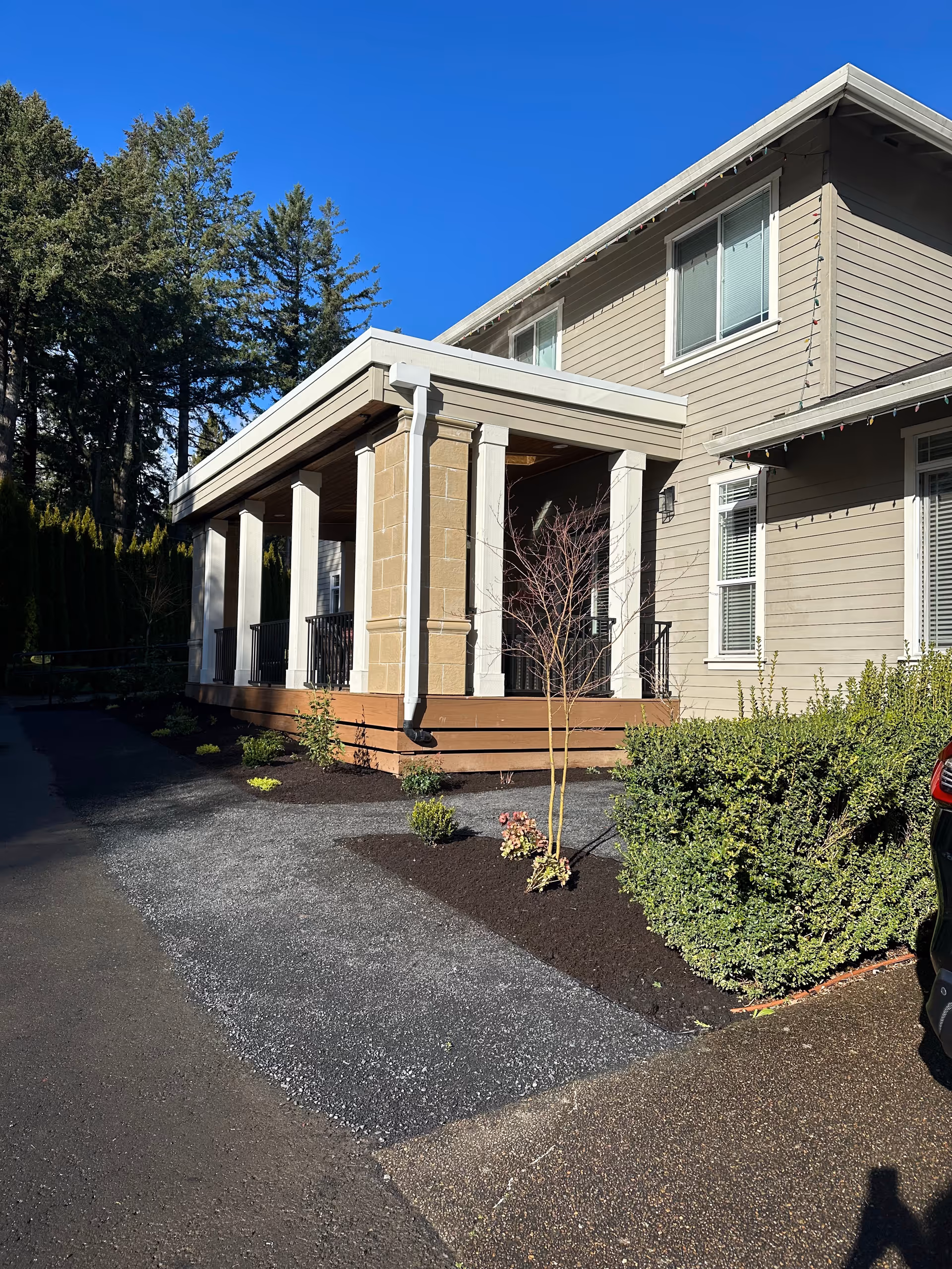 Exterior view of a two-story beige building with white trim and a covered porch supported by white columns. There are small landscaped plants and bushes along the driveway and walkway. The sky is clear and blue.