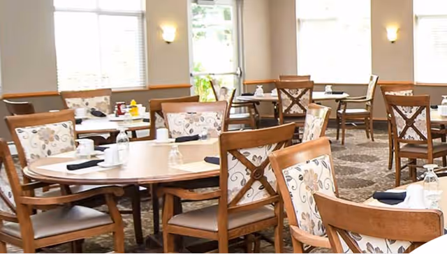A bright dining room with multiple round wooden tables and matching chairs featuring floral upholstery. Each table is set with white cups, saucers, napkins, and upside-down glasses. Large windows and a glass door allow natural light to fill the room.