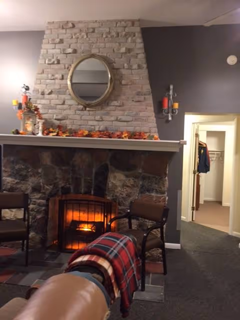 Cozy living room area with a stone fireplace featuring a lit fire. Above the fireplace is a brick chimney with a round mirror hanging on it. The mantle is decorated with autumn-themed items including leaves and candles. Two chairs are positioned near the fireplace, one with a red and black plaid blanket draped over it. The room has gray walls and an open doorway leading to another room with a closet visible.