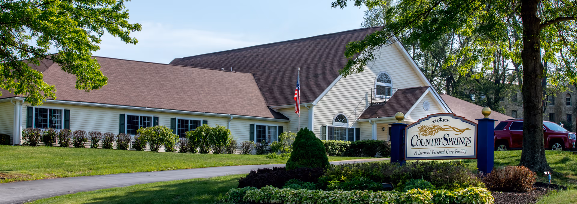 Exterior view of Country Springs, a licensed personal care facility, showing a single-story building with beige siding, brown roof, multiple windows, an American flag, and a landscaped lawn with bushes and trees.