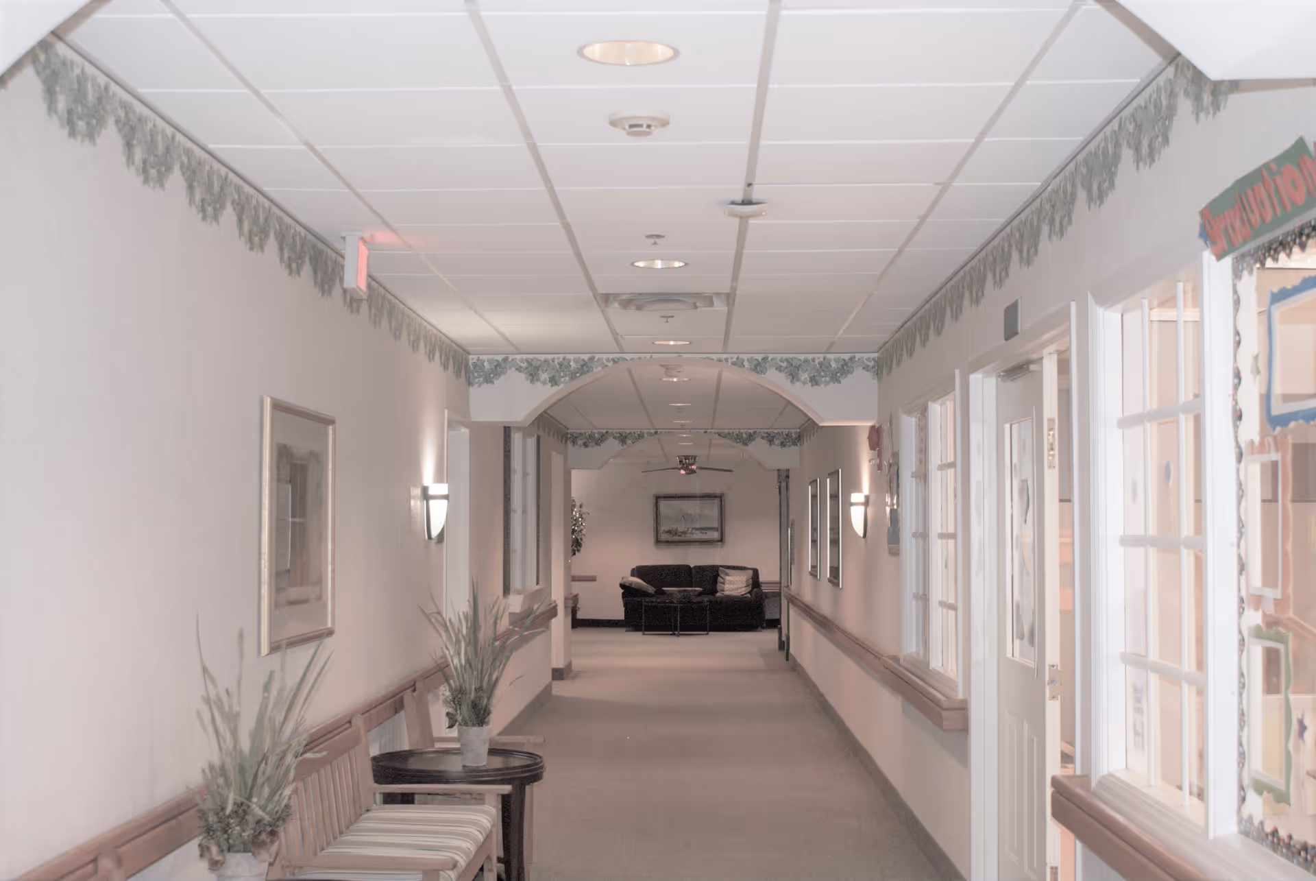 A long hallway in a senior living facility with beige walls and carpeted floor. There are wooden benches with striped cushions and potted plants on the left side. The ceiling has recessed lighting and decorative green leaf border near the top of the walls. At the end of the hallway, there is a black couch with pillows and a framed picture above it. On the right side, there are windows and a door with glass panes, and a colorful bulletin board partially visible.