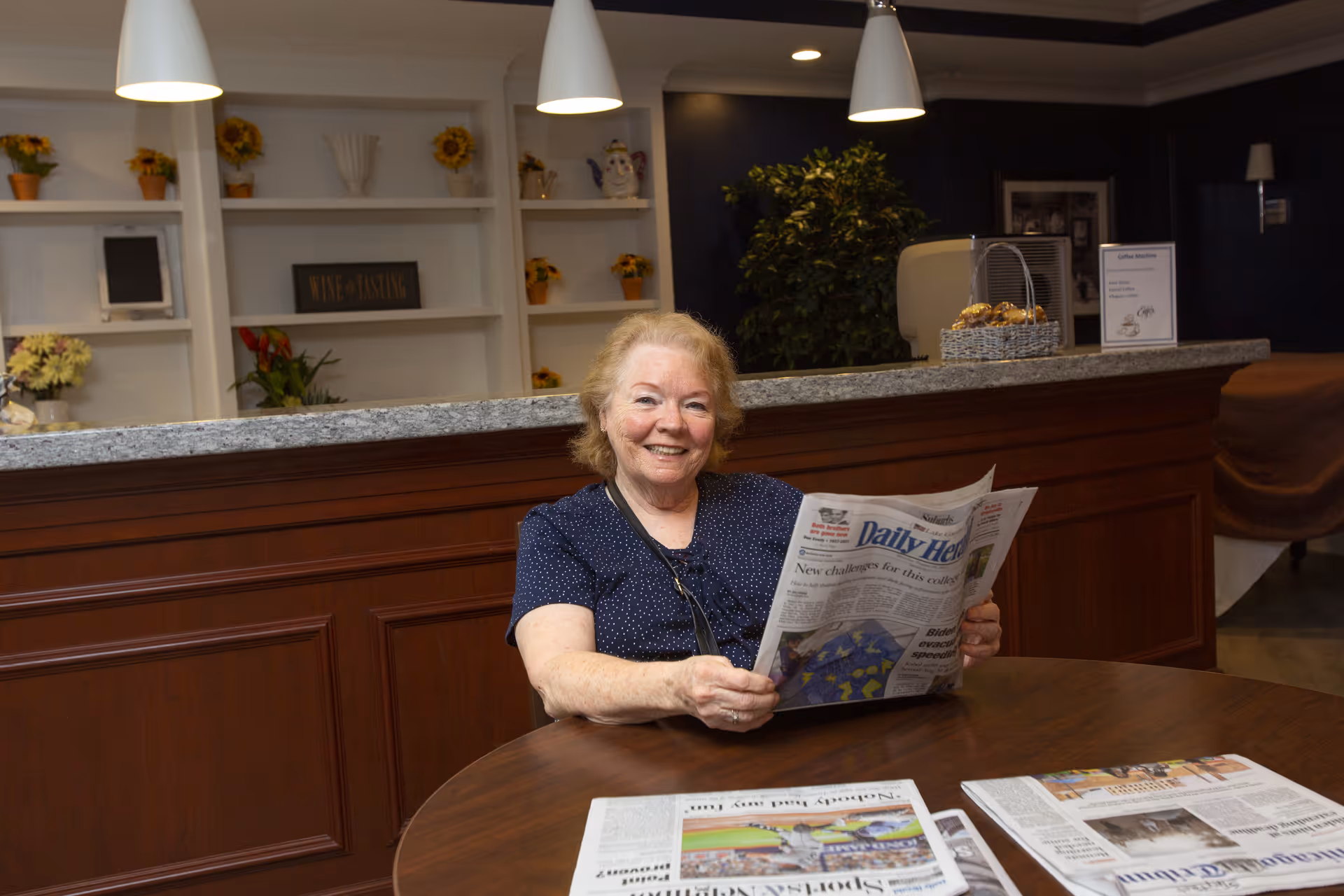 An elderly woman sitting at a round wooden table in a cozy interior space, smiling and holding a newspaper. Behind her is a counter with a granite top, shelves with decorative items and flowers, and pendant lights hanging from the ceiling.