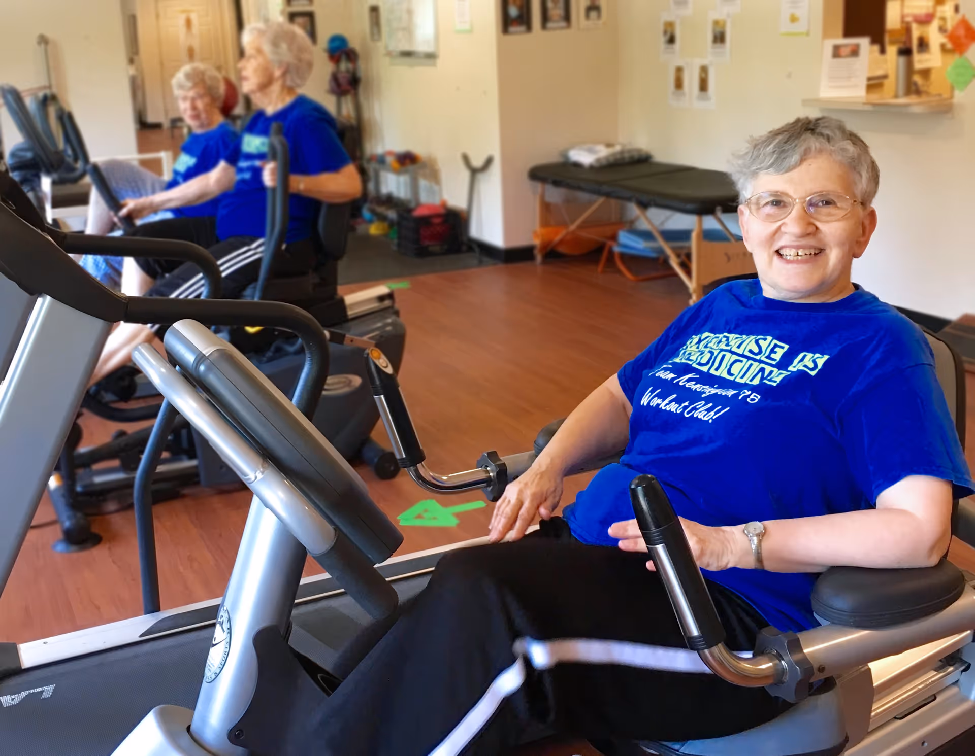 Three elderly women exercising on stationary recumbent bikes in a fitness room. The woman in the foreground is smiling at the camera, wearing glasses and a blue t-shirt. The room has wooden floors and exercise equipment, with some posters and a massage table in the background.