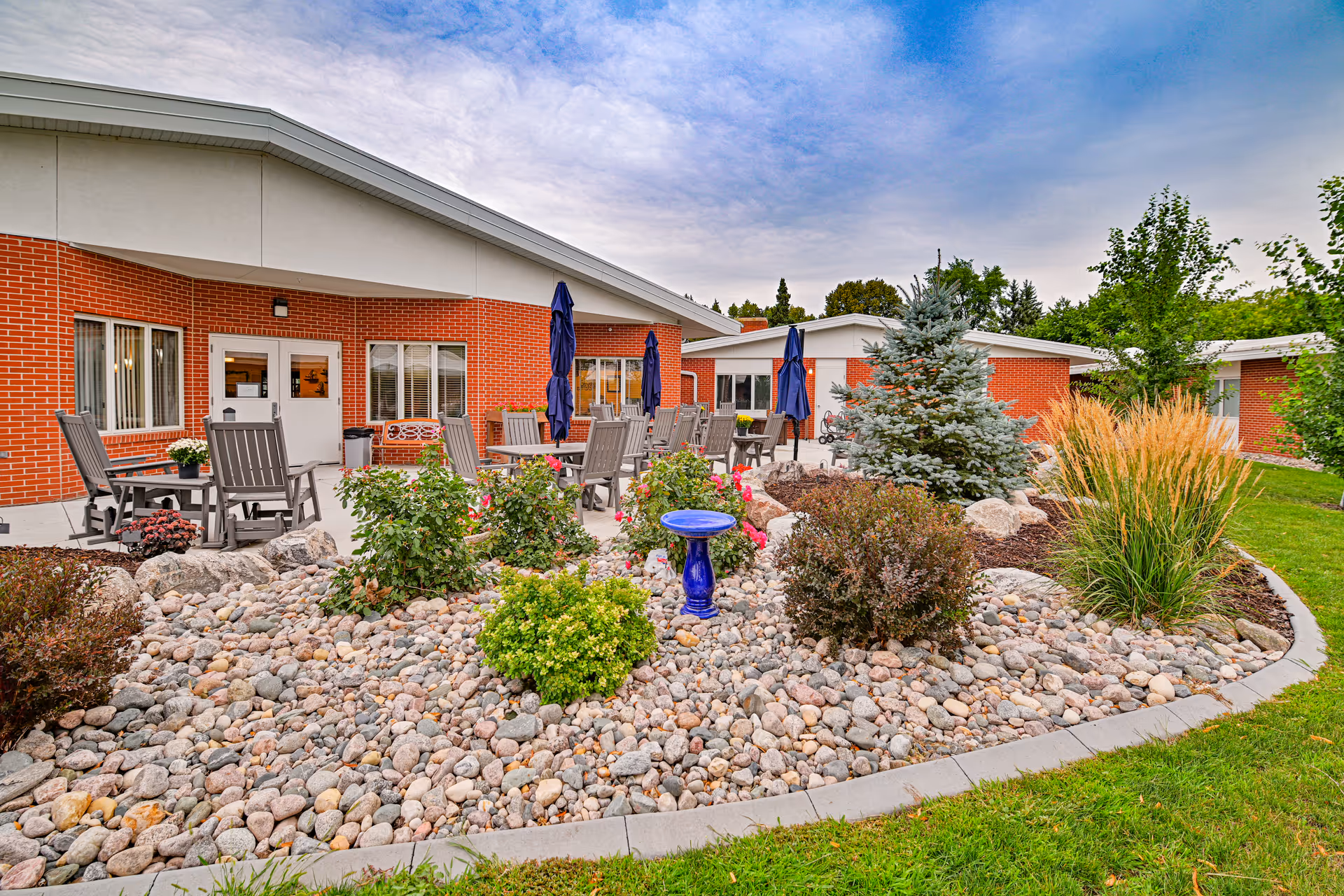 Outdoor patio area at Valley Senior Living on Cherry - Tufte Manor featuring multiple tables with chairs and closed umbrellas, surrounded by landscaped garden beds with rocks, shrubs, flowers, and a small blue birdbath. The building has red brick walls and white trim under a partly cloudy sky.