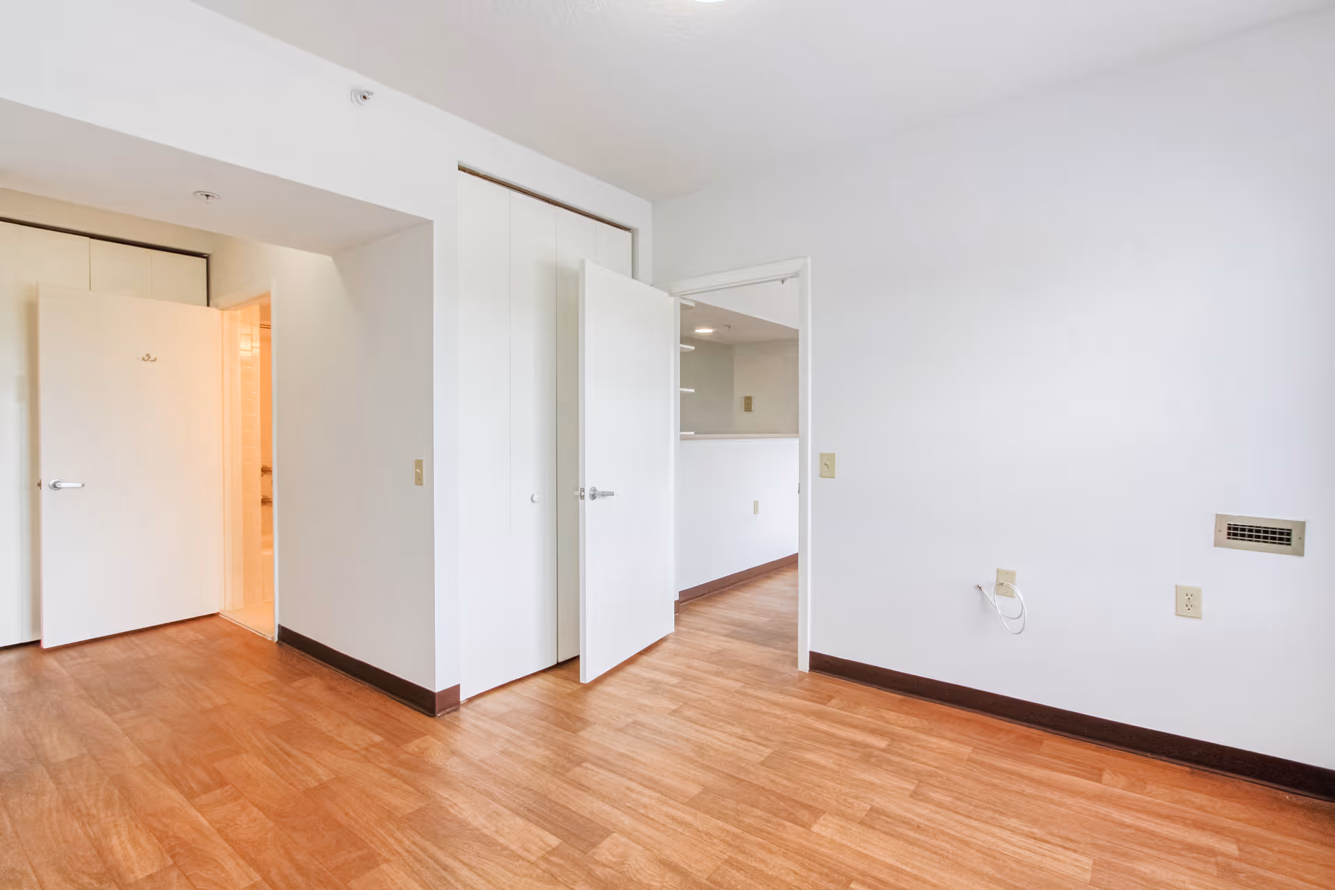 Empty room with wood flooring, white walls, and two open doors showing a closet and a hallway leading to another room.