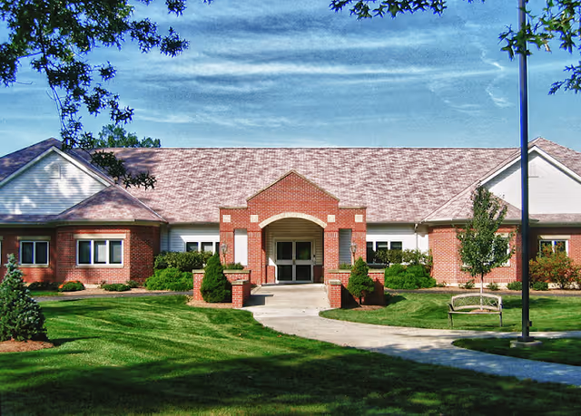 Front exterior view of a single-story brick building with a peaked roof, an arched entrance, surrounded by well-maintained green lawn, shrubs, trees, a bench, and a lamppost under a partly cloudy blue sky.